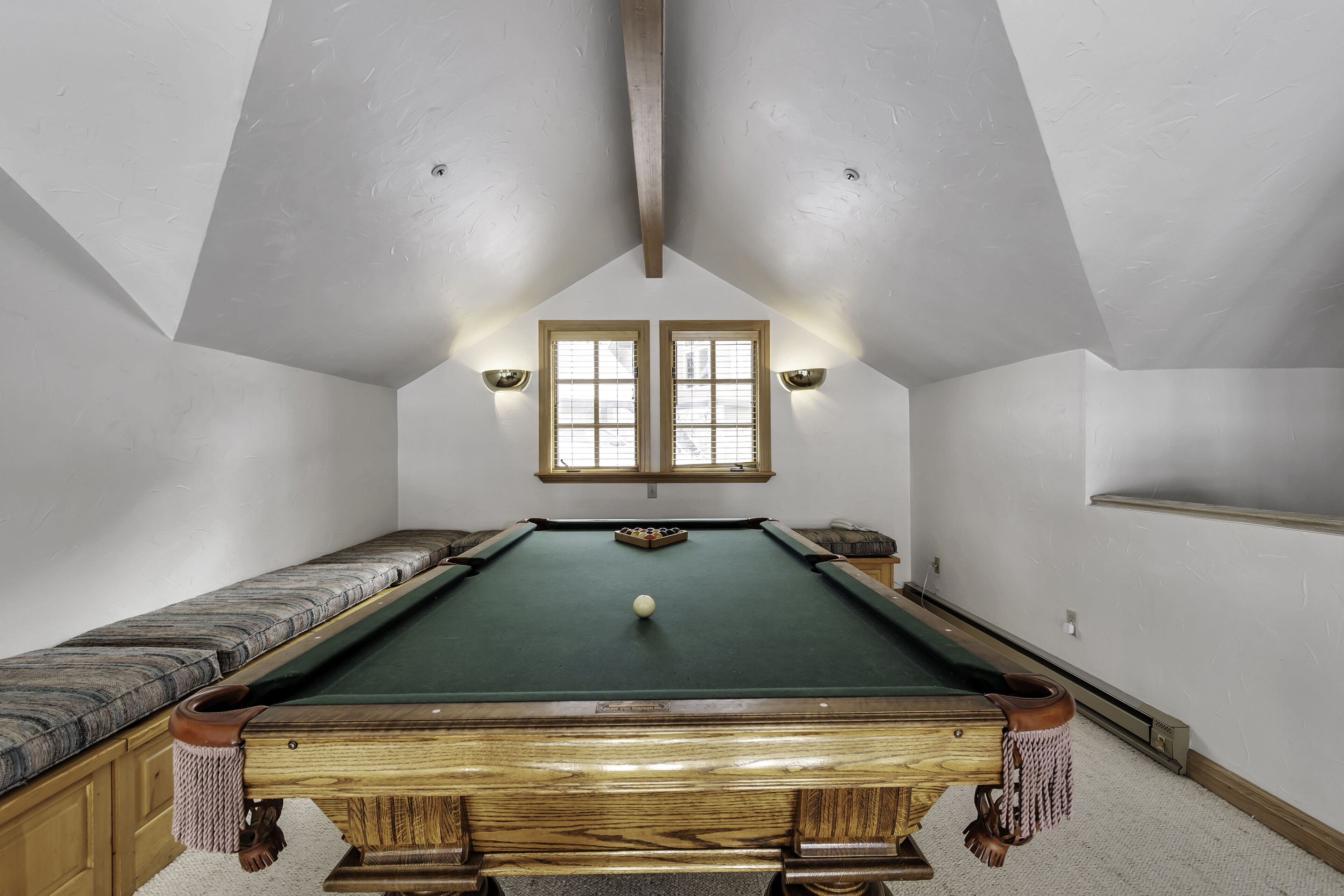 A pool table with a ball, wooden benches, and two windows with light fixtures.