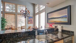 Eine moderne Küche mit einer Granitabdeckung, einer Edelstahlspüle und einer Kristallleuchter. Es gibt große Fenster mit Jalousien, einen Essbereich mit Stühlen und ein Bild an der Wand.
