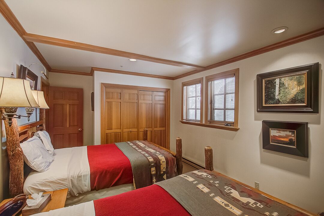 A hotel room with two beds, wooden wardrobe, framed pictures on the wall, and a window with blinds.