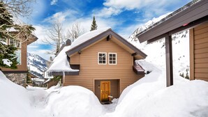 Ein mit Schnee bedecktes Holzhaus mit einer Haustür, umgeben von Schneewehen und Bergen im Hintergrund.