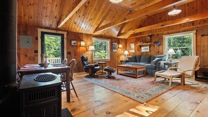 A cozy cabin interior with wooden walls, a fireplace, a dining area, and a living room with a sofa and chairs.