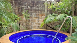 Un jacuzzi rond, de couleur bleue à l’intérieur, bordé d’une structure beige, installé contre un mur de pierre agrémenté de verdure.