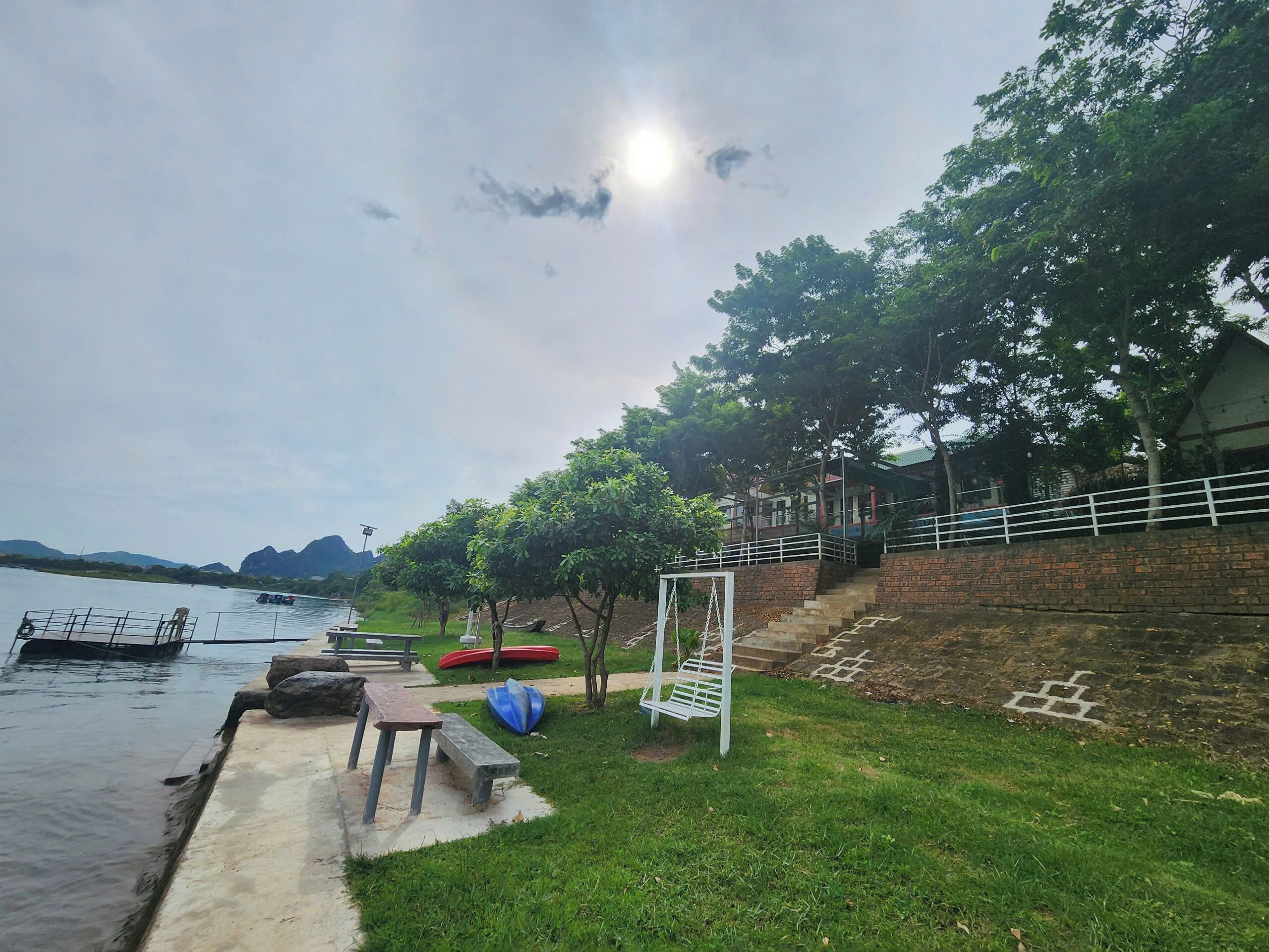 A riverside park with a concrete walkway, picnic tables, a swing set, and boats on the water.