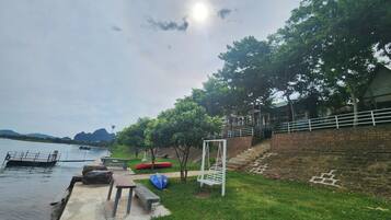 A riverside park with a concrete walkway, picnic tables, a swing set, and boats on the water.