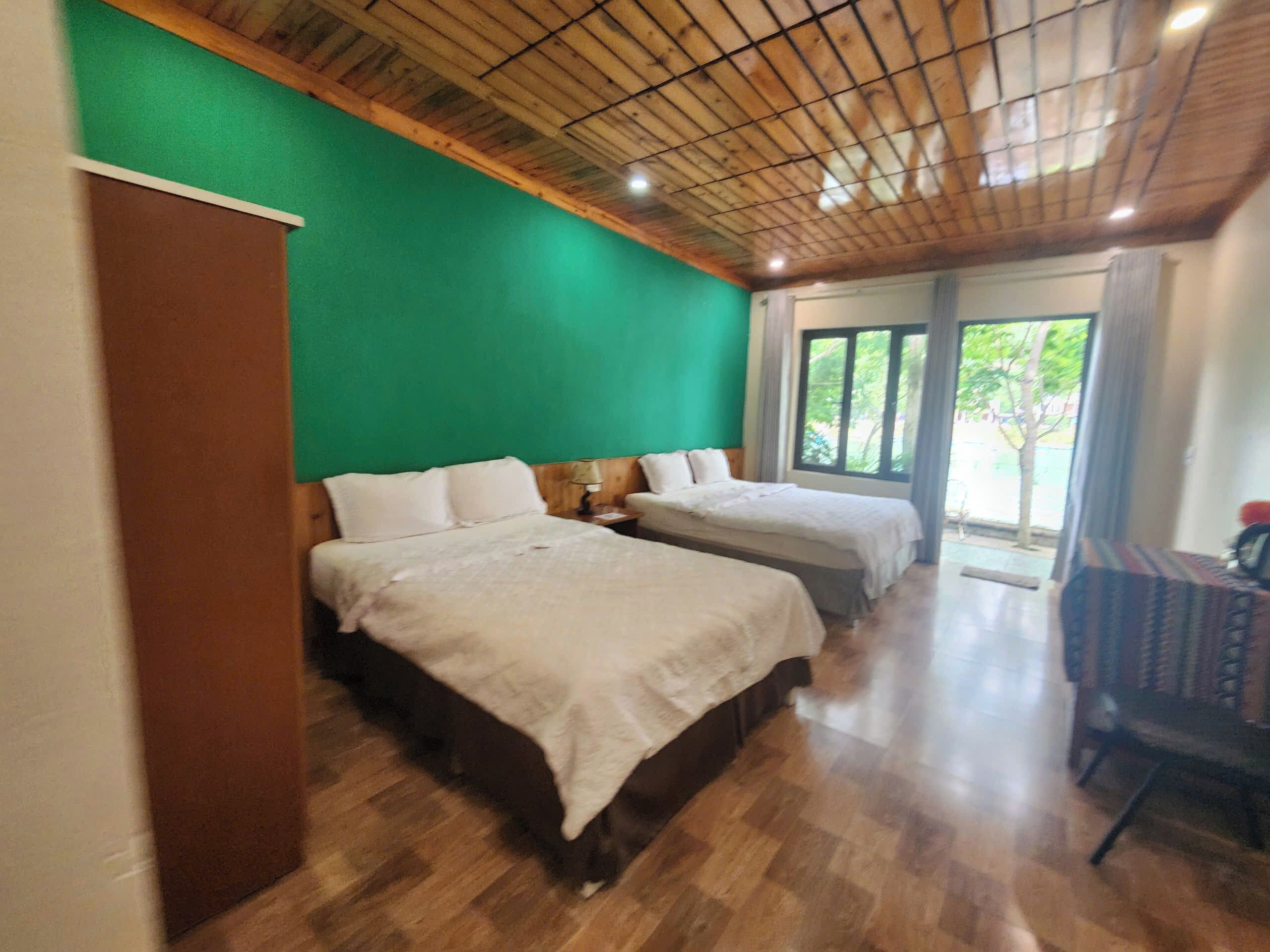 Een kamer met twee bedden, houten vloer, een groene wand en een groot raam met uitzicht op groen.