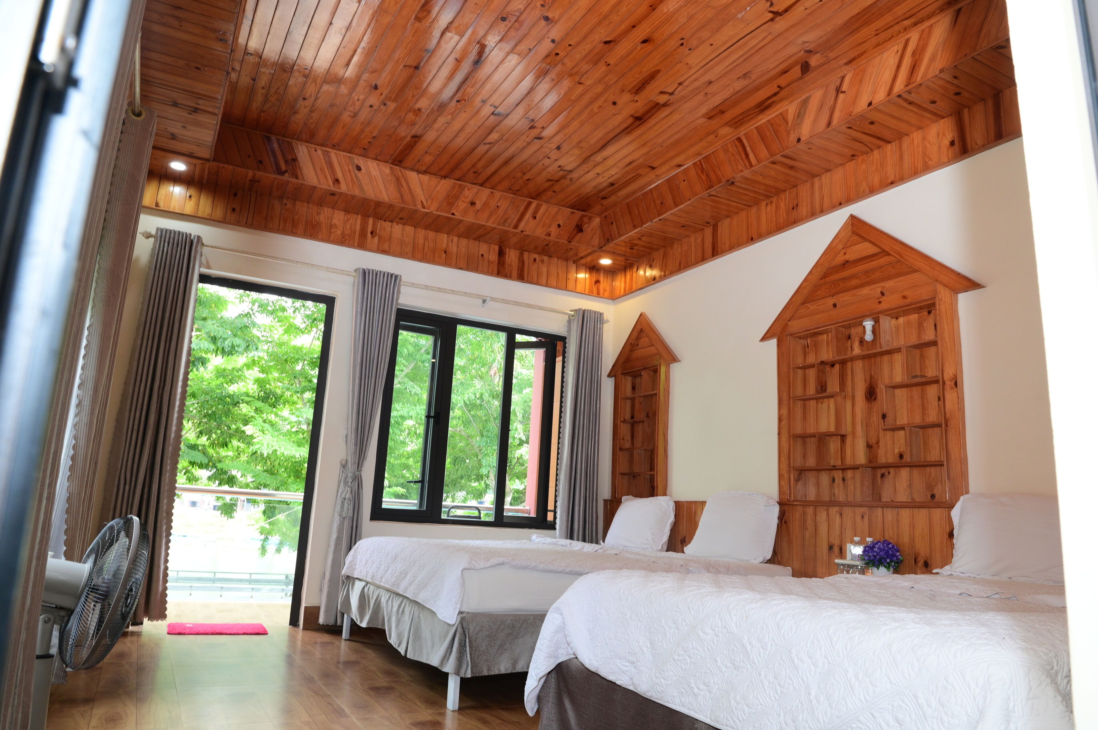 A bedroom with two beds, wooden ceiling, large windows, and a view of greenery outside.