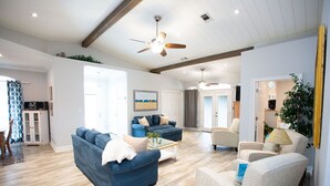 A spacious living room with a blue sofa, a white armchair, and a coffee table. There is a ceiling fan, a painting on the wall, and a door leading to another room.