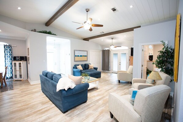 A spacious living room with a blue sofa, a white armchair, and a coffee table. There is a ceiling fan, a painting on the wall, and a door leading to another room.