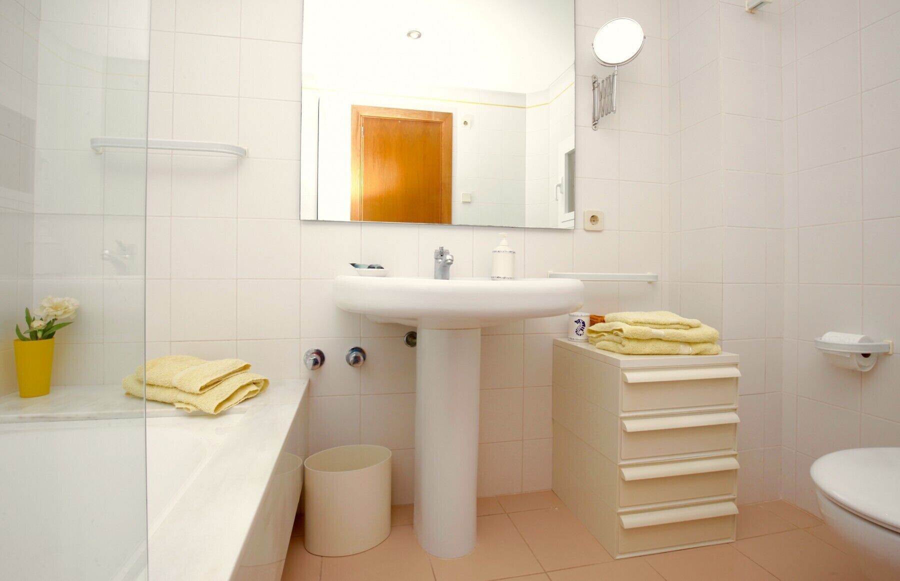 Et badeværelse med en hvid vask, spejl og toilet. Der er en håndklæderække med håndklæder og en skraldespand.