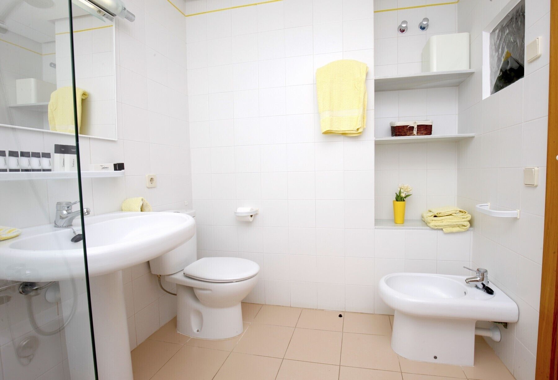 A bathroom with a toilet, sink, and bidet. There are shelves with toiletries and a yellow towel.