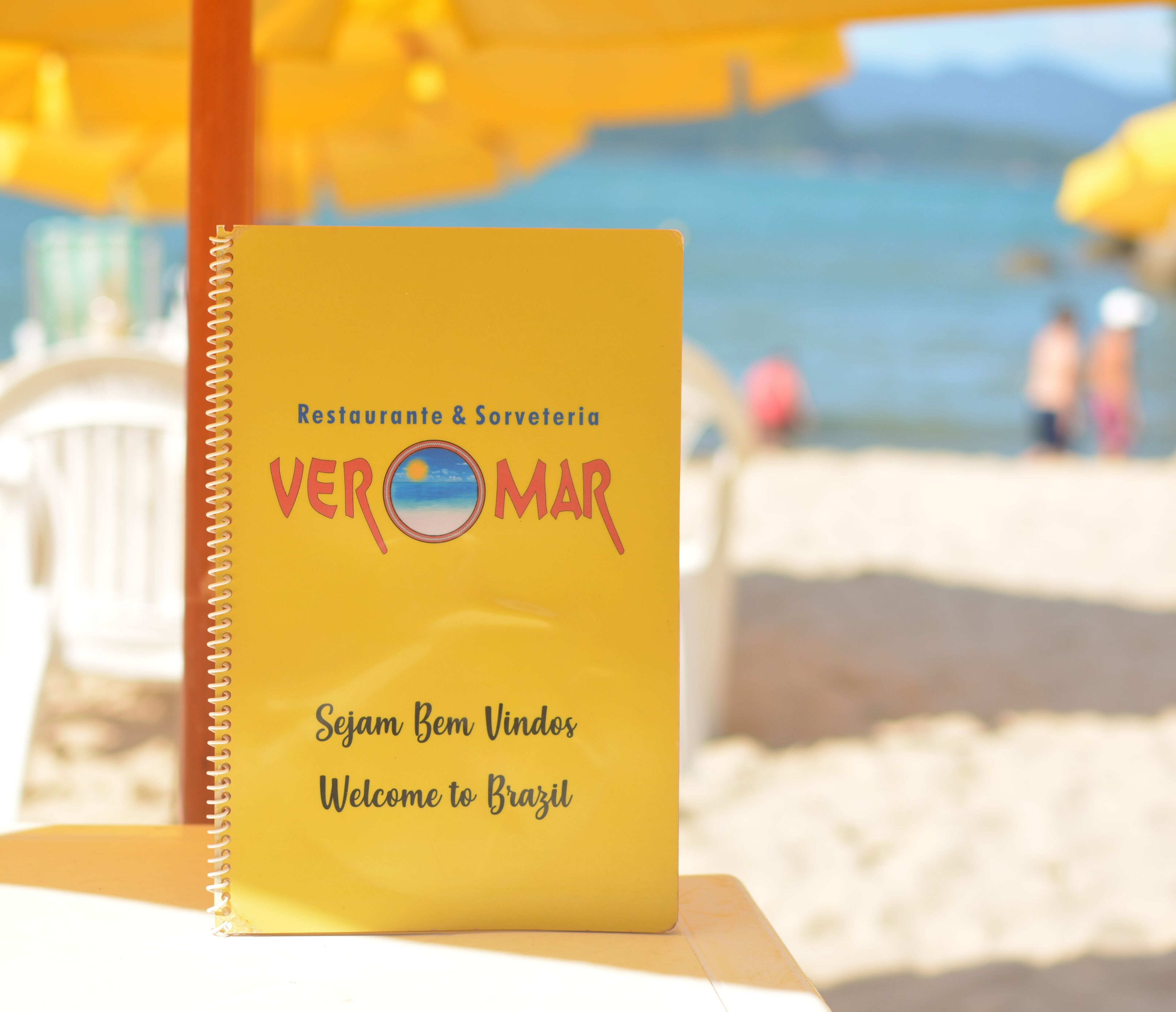 Una libreta en espiral amarilla con el título 'VEROMAR' y la inscripción 'Sejam Bem Vindos Welcome to Brazil' en un entorno playero.