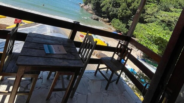 A wooden deck with a table and chairs overlooking a beach with clear blue water and distant mountains. - Pousada Veromar (Paraty)