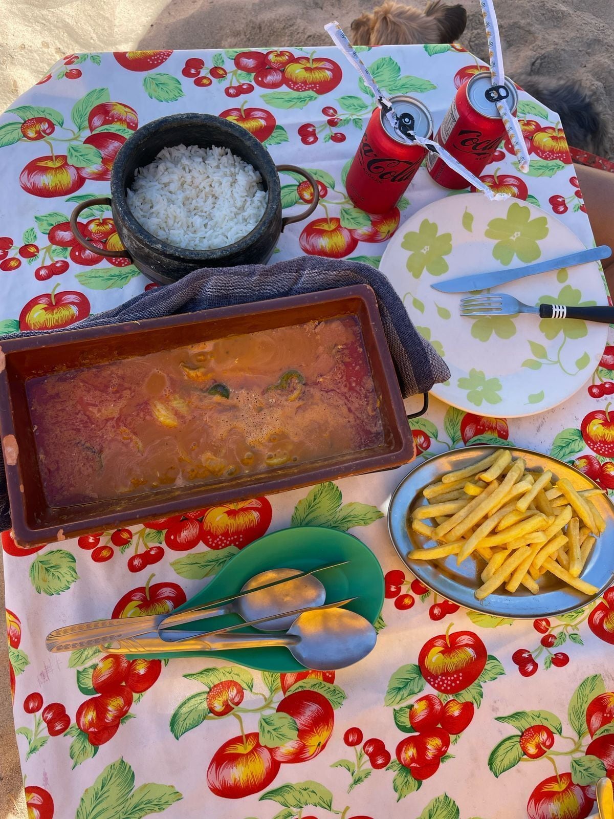A table set with a fruit-themed tablecloth, a bowl of rice, a dish with a red sauce, a plate of fries, a can of soda, and cutlery.