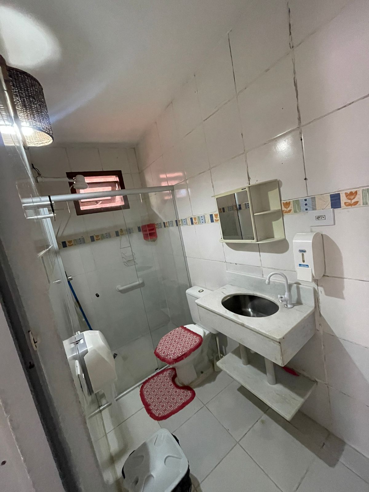 A small, compact bathroom with a toilet, bidet, and sink. There is a window with a red mat and a shower area with a glass door.