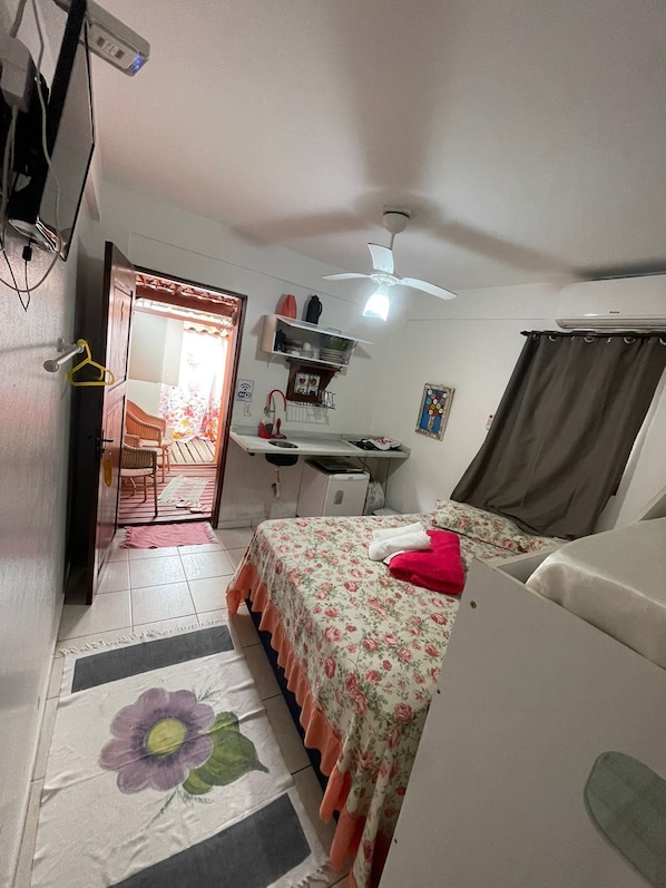 Una habitación compacta con cama, escritorio y silla. Hay un ventilador de techo y una ventana con cortinas.