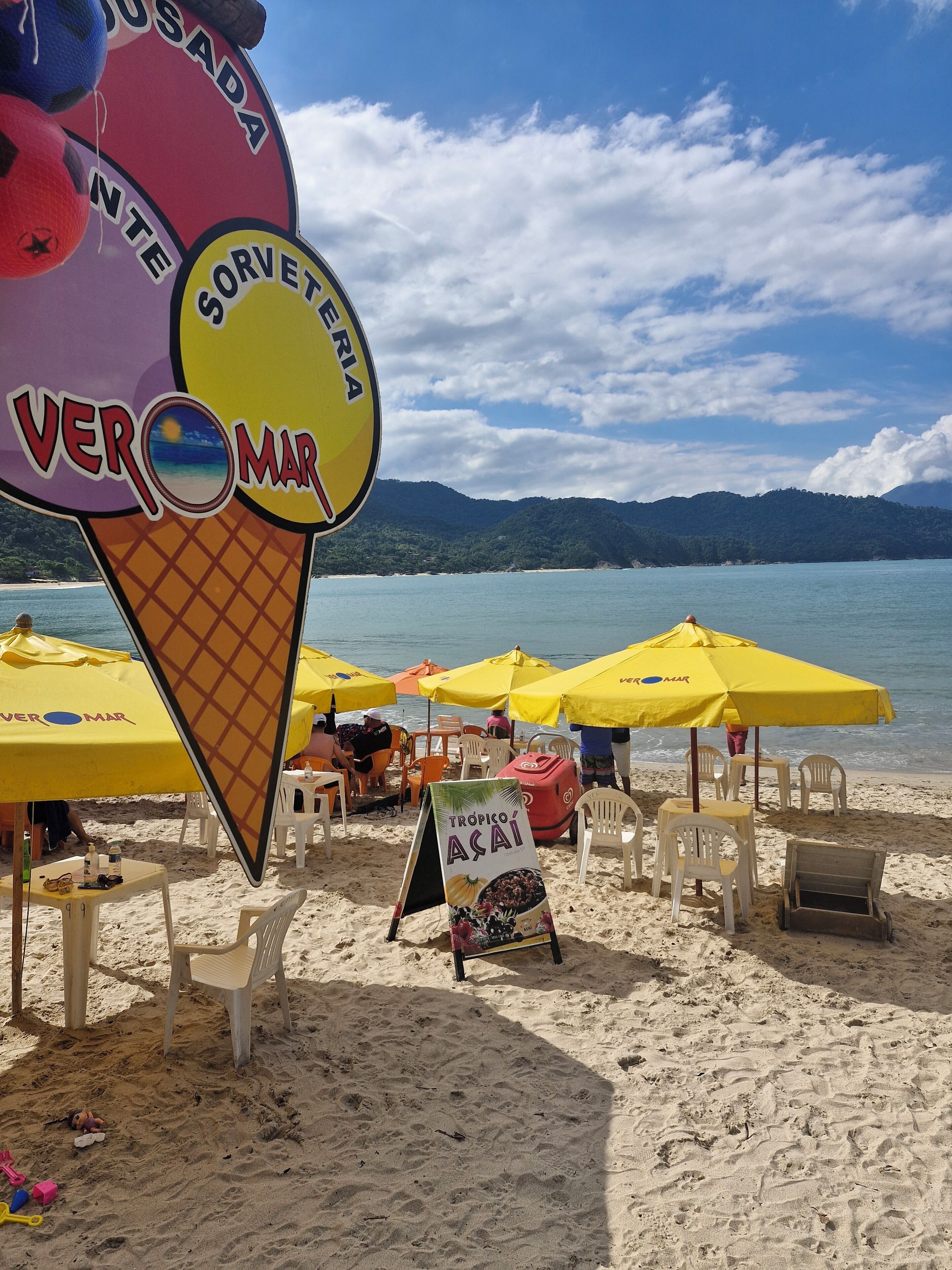 A beach with yellow umbrellas, a sign for 'Tropico Açaí', and a large ice cream cone sign.