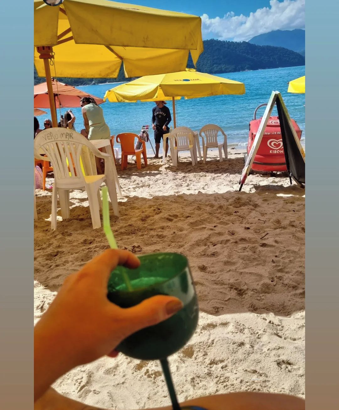 A beach scene with yellow umbrellas, a lifeguard tower, and a person holding a drink.