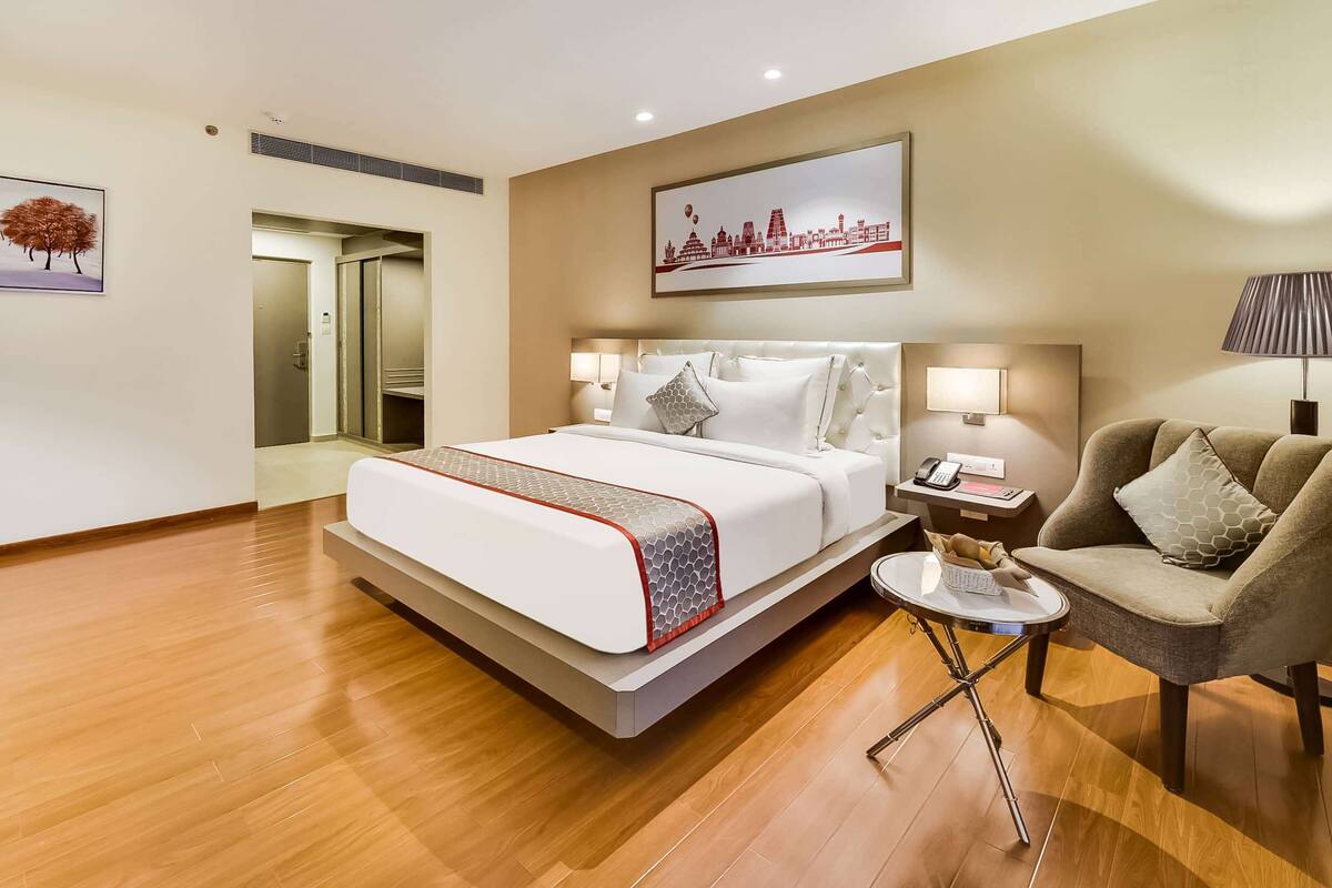 Ein Hotelzimmer mit einem großen Bett, Nachttischlampen, einem kleinen Tisch, einem Sessel und einem Stadtbild an der Wand.