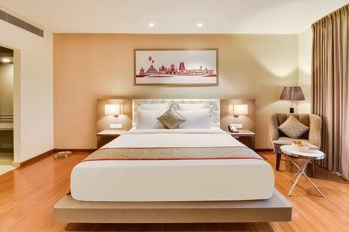Ein Hotelzimmer mit einem großen Bett, zwei Nachttischen mit Lampen, einem Sessel und einem Stadtbild an der Wand.