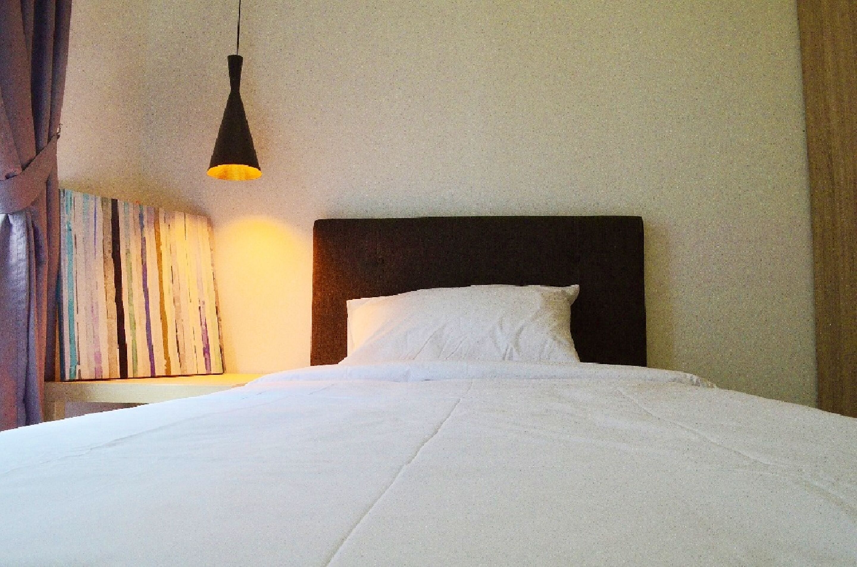 A neatly made bed with white linens, a dark headboard, and a single hanging lamp providing light.