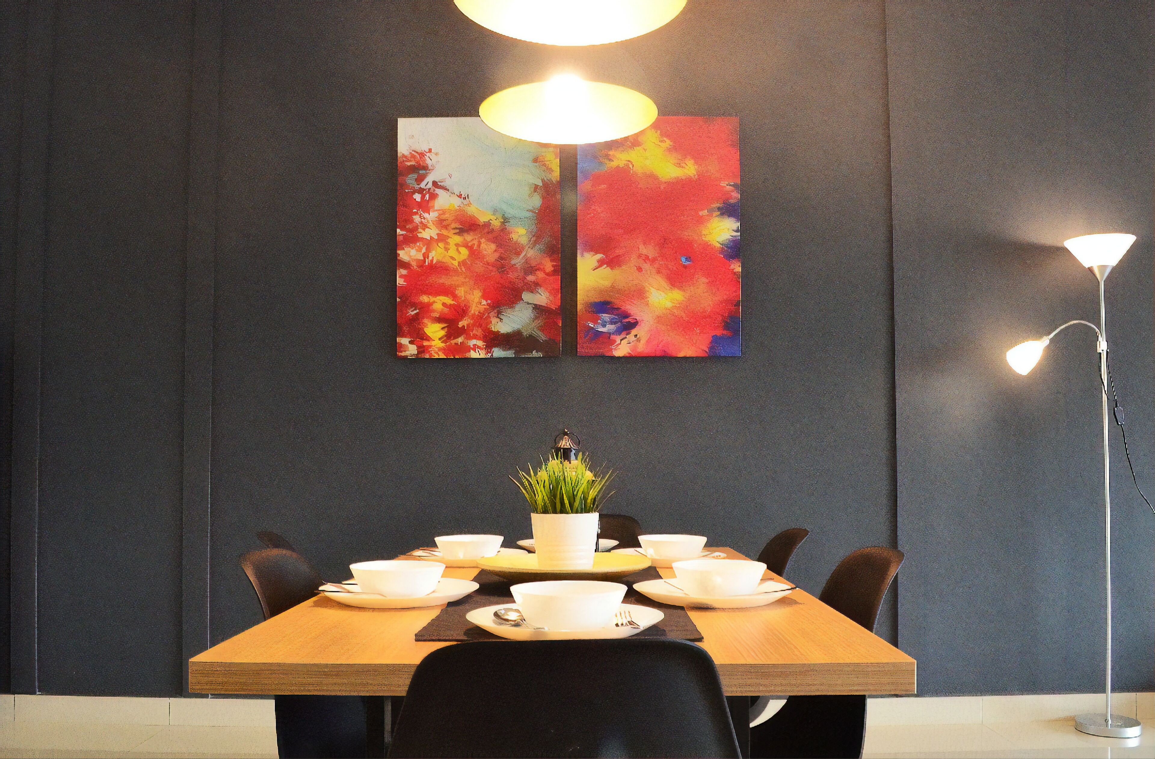 Un comedor con una mesa de madera dispuesta para cuatro personas, dos cuadros abstractos en la pared, una planta en maceta y una lámpara de pie.