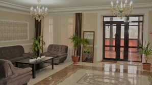 A spacious living room with a brown sofa set, a coffee table, and two potted plants. There is a chandelier hanging from the ceiling and a large mirror on the wall.