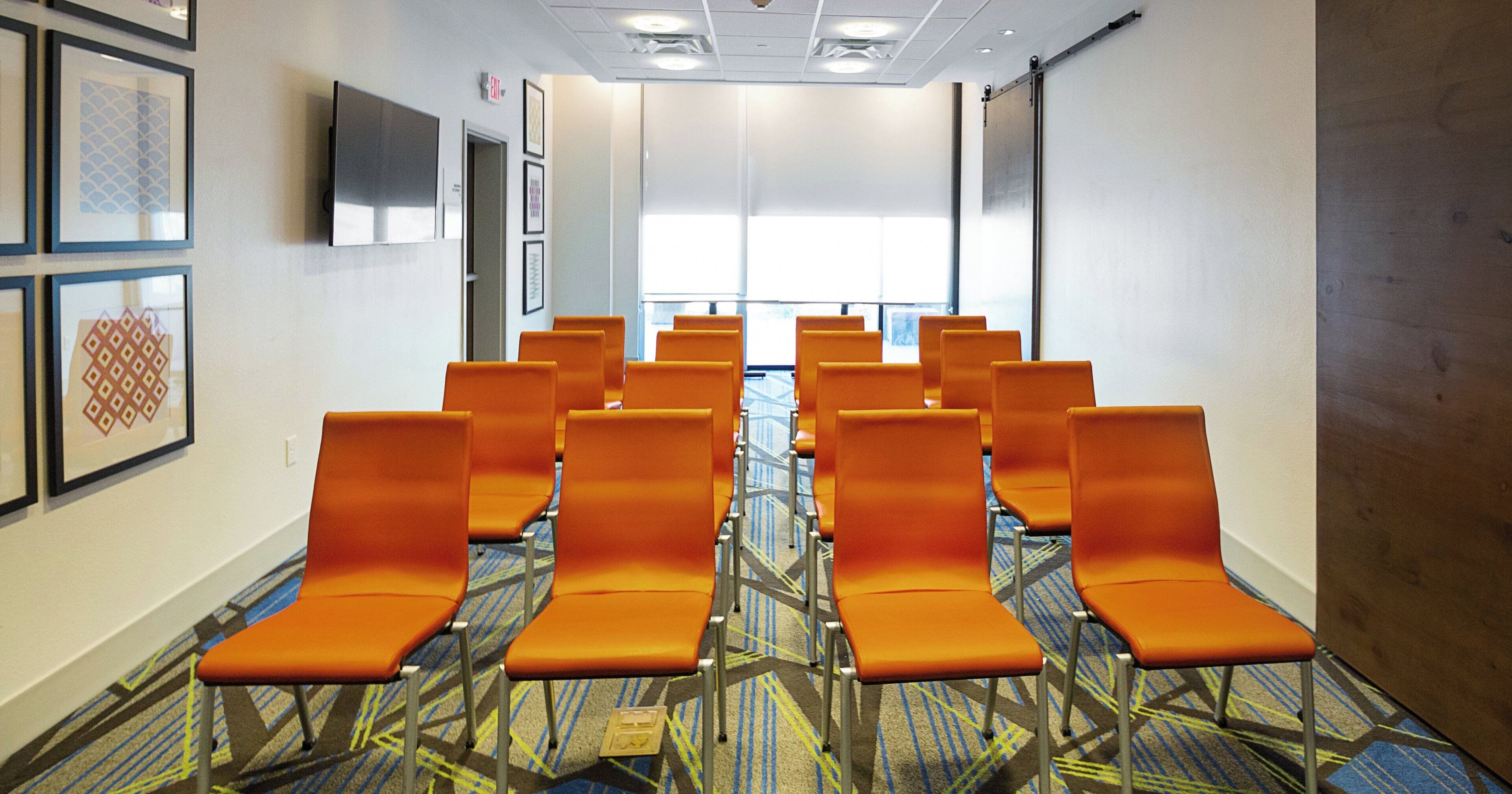 A hotel lobby with rows of orange chairs, a flat-screen TV mounted on the wall, and patterned carpet.