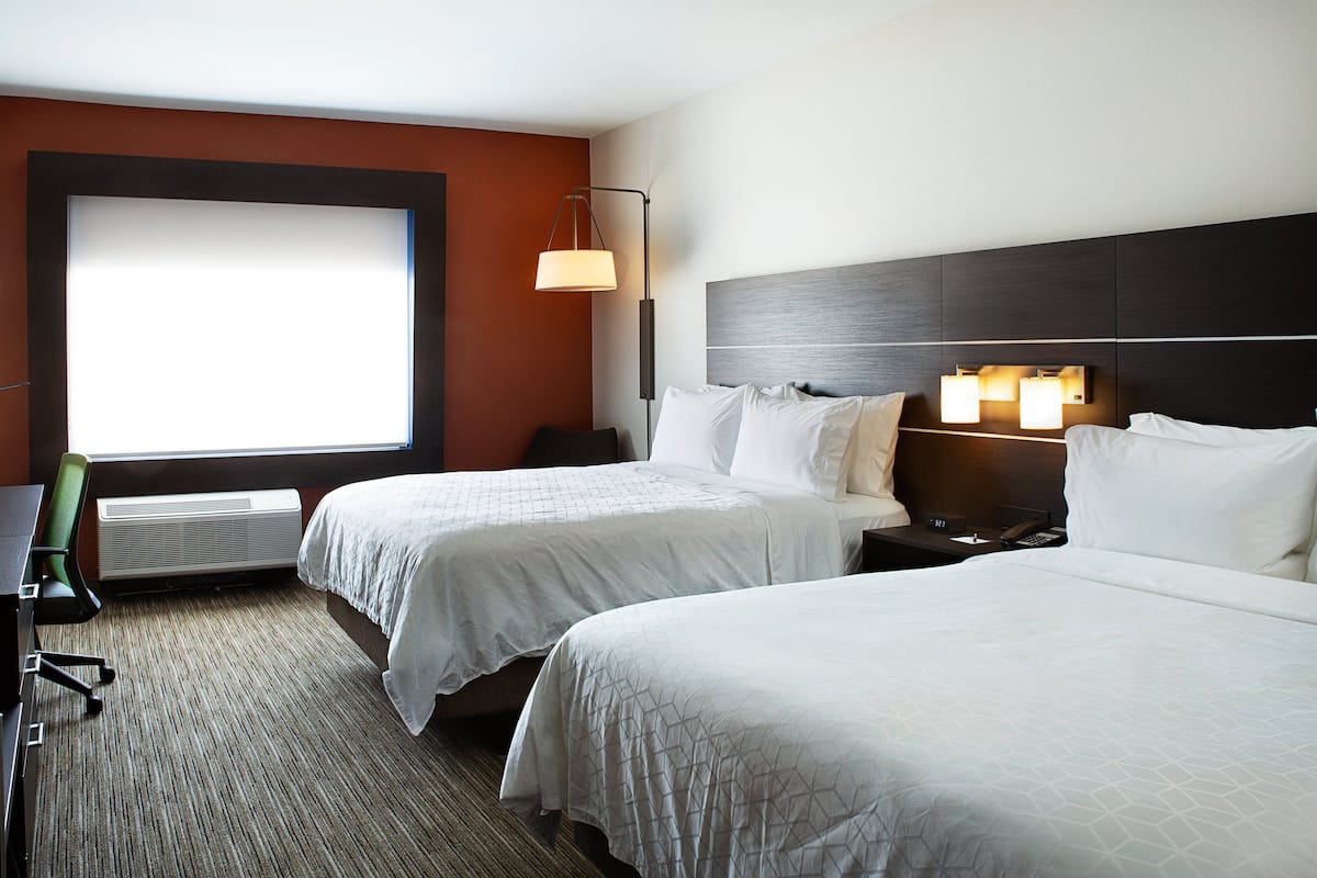 Habitación de hotel con dos camas, un escritorio, una silla, una ventana con persianas, una lámpara de pared y un termostato.