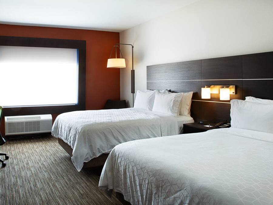 Habitación de hotel con dos camas, un escritorio, una silla, una ventana con persianas, una lámpara de pared y un termostato.