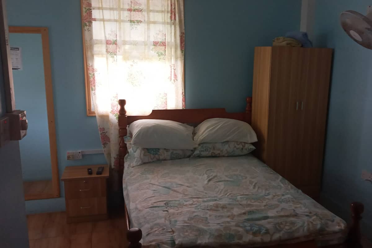 Ein Schlafzimmer mit einem hölzernen Bett, einem Kleiderschrank, einem Fenster mit blumigen Vorhängen und einem Wandventilator.