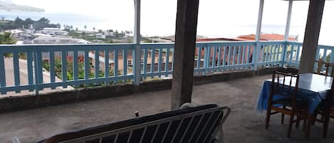 A covered outdoor area with a table and chairs, a metal railing, and a view of buildings and water in the distance.