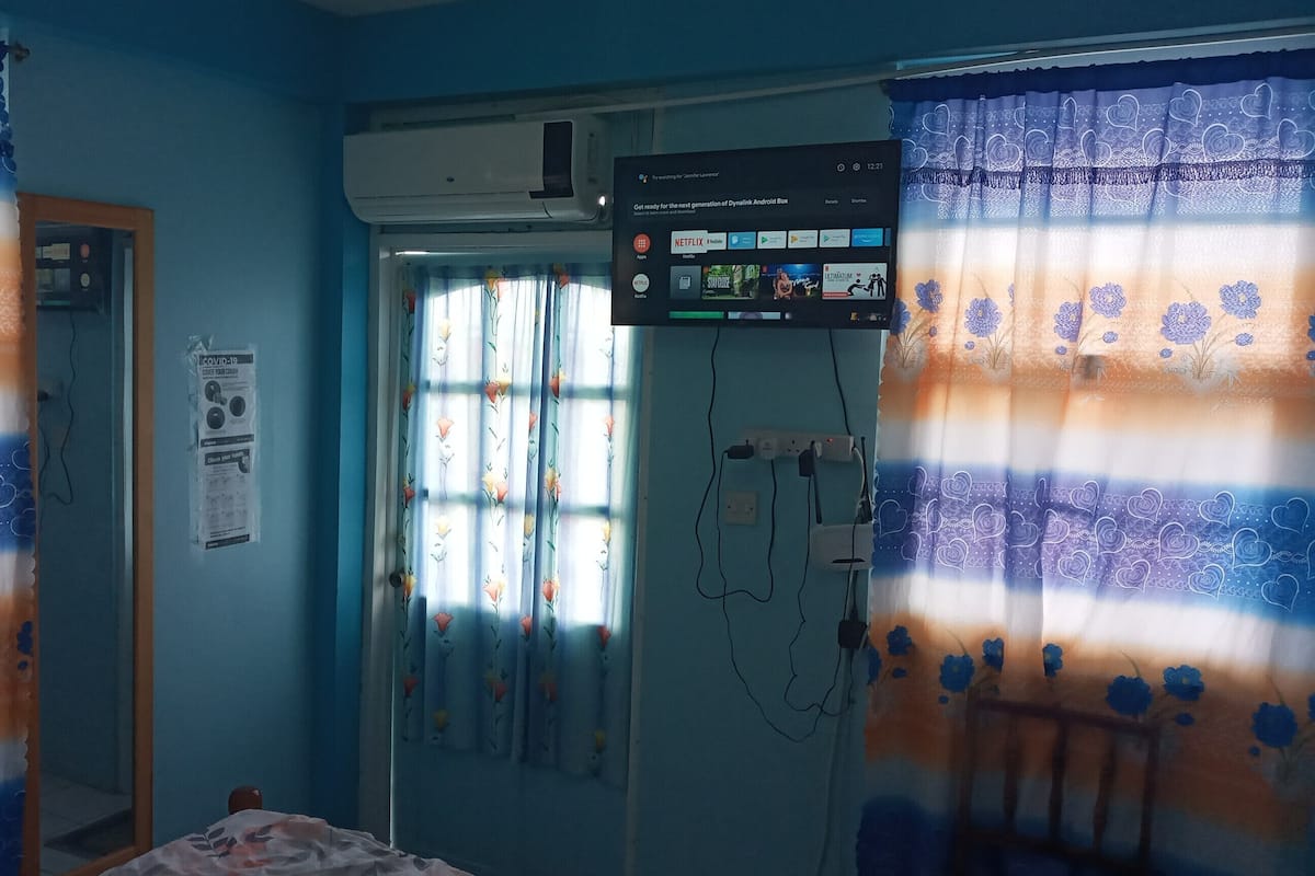 Ein Zimmer mit einem Bett, einem Fenster mit floralem Vorhang, einer Klimaanlage und einem an der Wand befestigten Fernseher, der eine Netflix-Oberfläche zeigt.