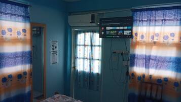 Une chambre avec un lit, une fenêtre ornée de rideaux à motifs floraux, un appareil de climatisation et un téléviseur fixé au mur affichant l’interface de Netflix.