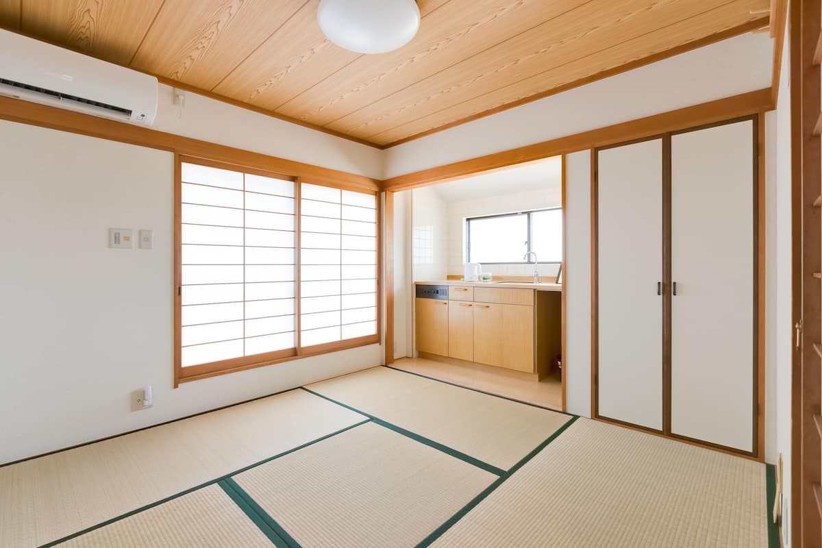 A traditional Japanese room with tatami flooring, sliding doors, and a kitchen area visible through a window.