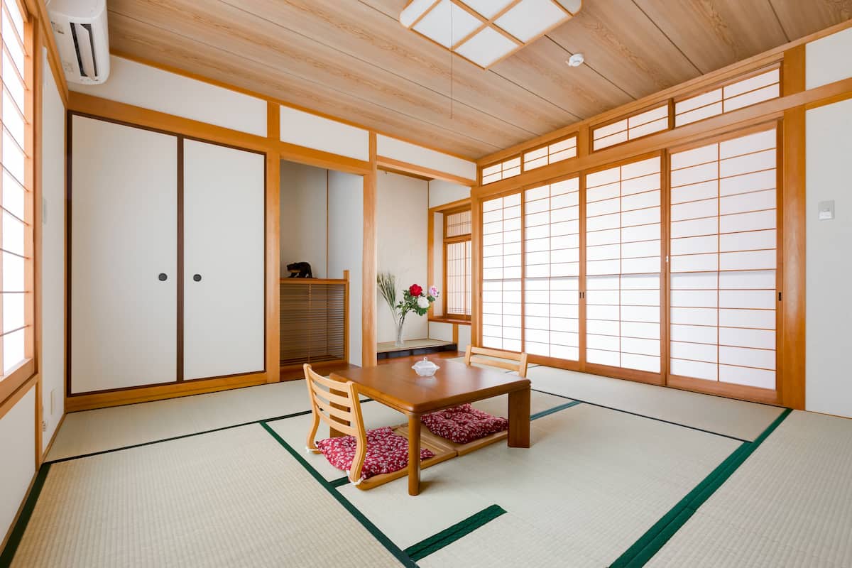 A traditional Japanese room with tatami flooring, a low wooden table, a chair, sliding doors, and a large window with shoji screens.