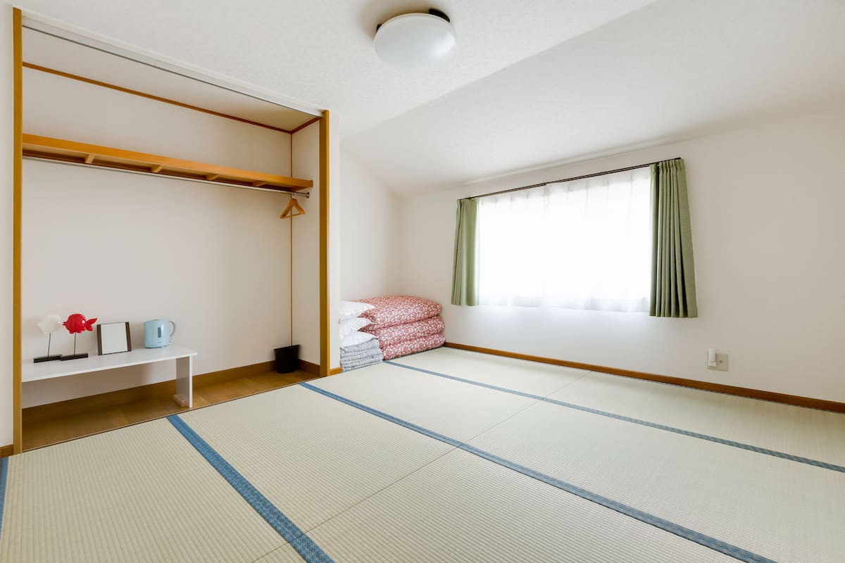 A room with tatami flooring, a wooden shelf with a kettle, pillows, and a framed picture.