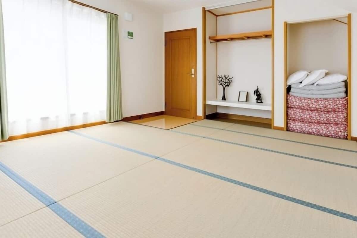 A room with tatami flooring, a wooden door, a shelf with pillows and a small statue, and a window with green curtains.