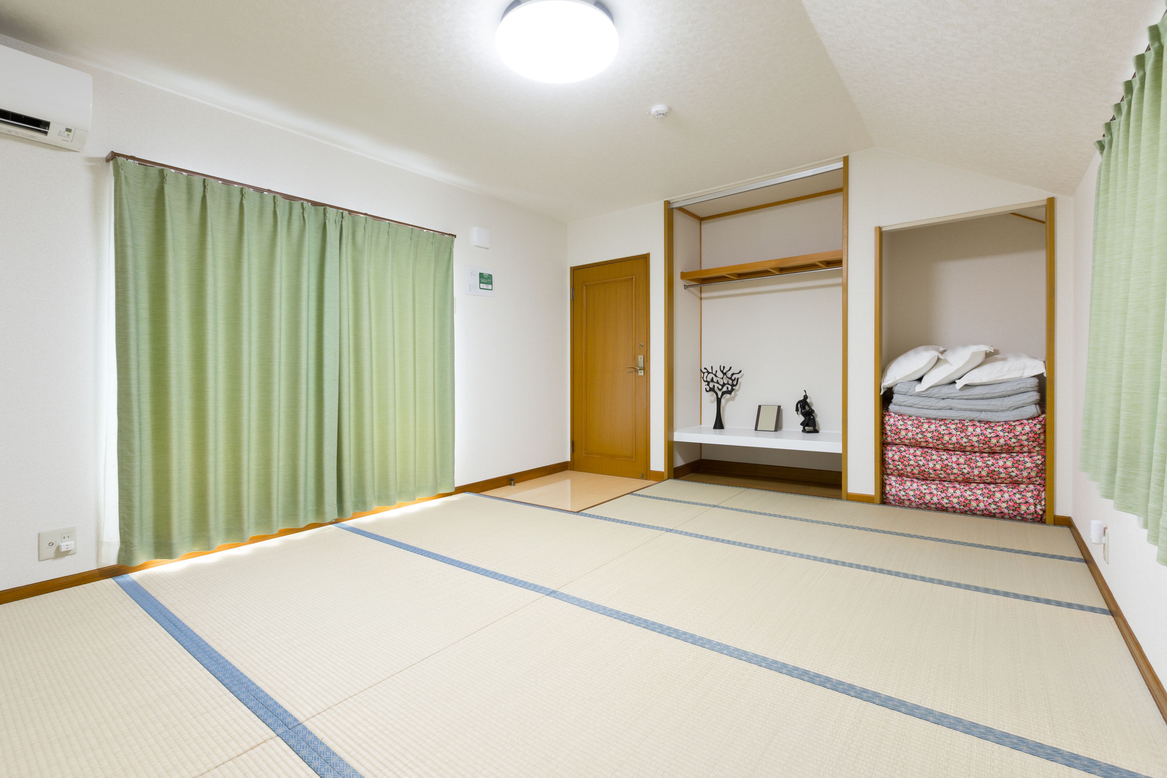 Ein Zimmer mit Tatami-Böden, einem Holzschrank, einem Regal mit Kissen und einem grünen Vorhang.