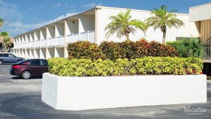 Un hôtel à la façade blanche, doté d’un espace extérieur couvert avec une rangée de balustrades blanches, et d’un jardin aménagé avec des palmiers et des arbustes.
