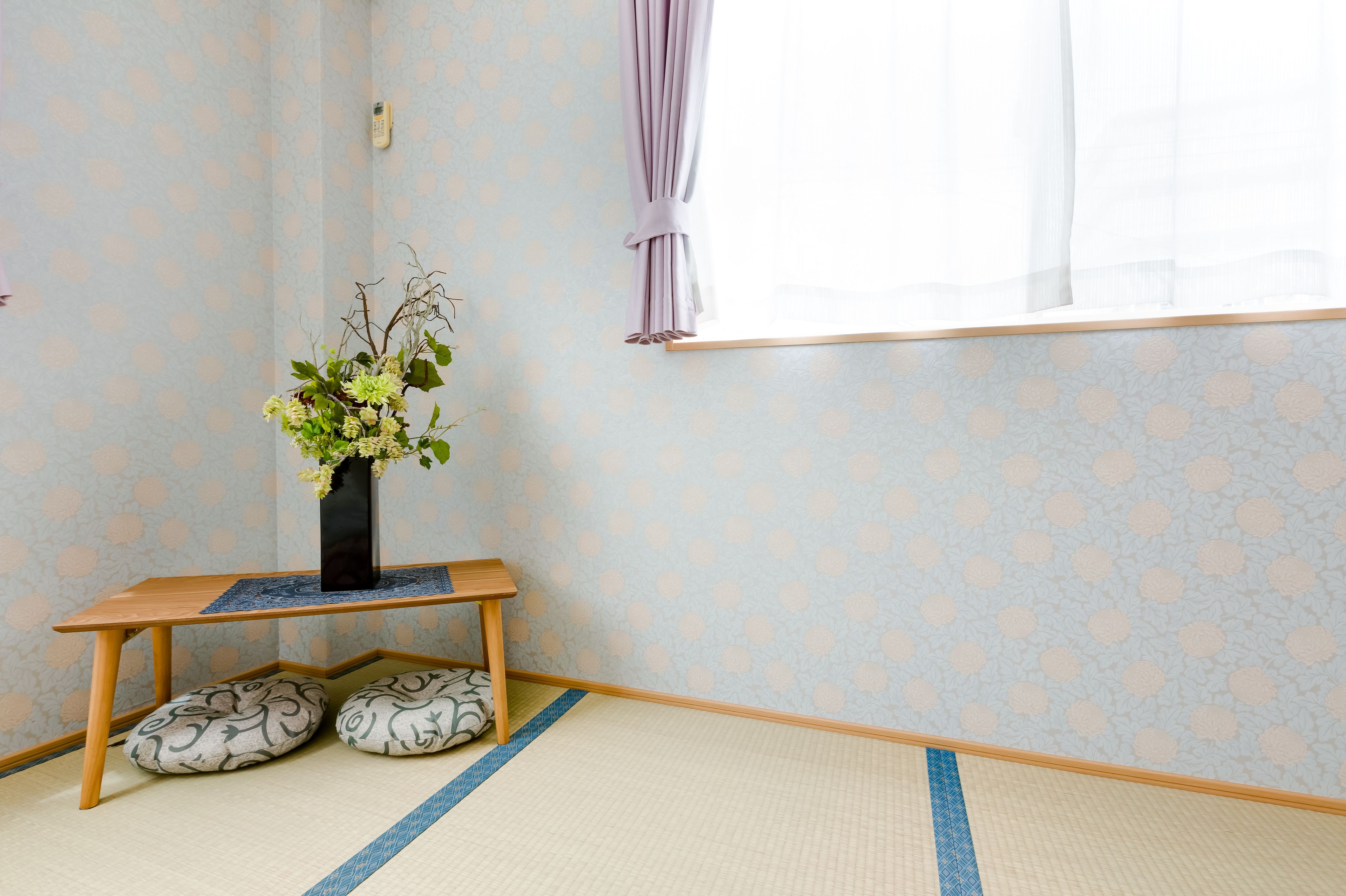 Um quarto com piso de tatame, um banco de madeira, um vaso com flores e uma janela com cortina.