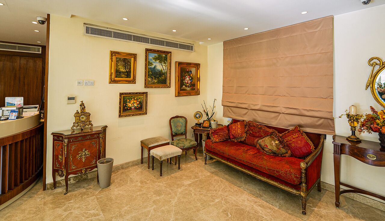 A traditional room with a red sofa, a wooden cabinet, a small table, and a chair. There are framed pictures on the wall and a mirror on the right.