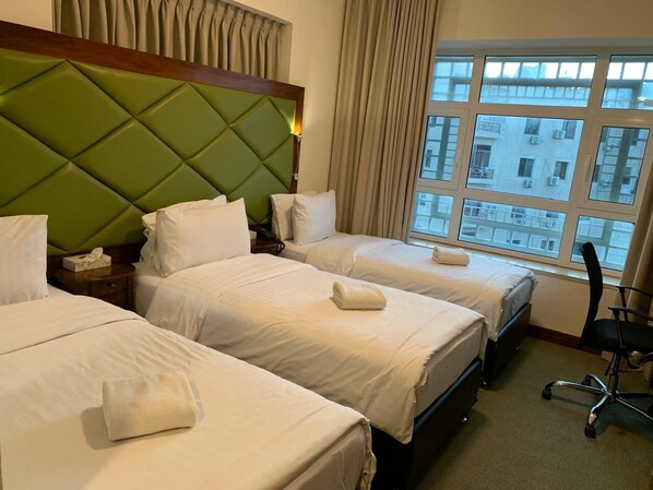 A hotel room with three beds, a green headboard, a large window with a city view, and a black office chair.