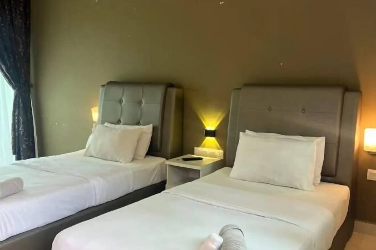 A hotel room with two beds, each with a headboard and pillow. There is a nightstand with a lamp between the beds.