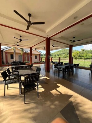 Un espace de restauration extérieur abrité, doté de plusieurs tables et chaises, de ventilateurs de plafond et offrant une vue sur un paysage verdoyant.