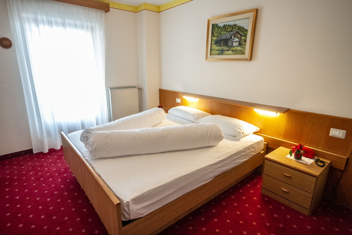 A hotel room with a wooden bed, white linens, a nightstand with a lamp, a painting on the wall, and a window with curtains.
