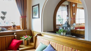 A cozy living room with a striped sofa, a red cushion, a small table with a plant, and a window with curtains. - Hotel Stella Alpina (Predazzo)