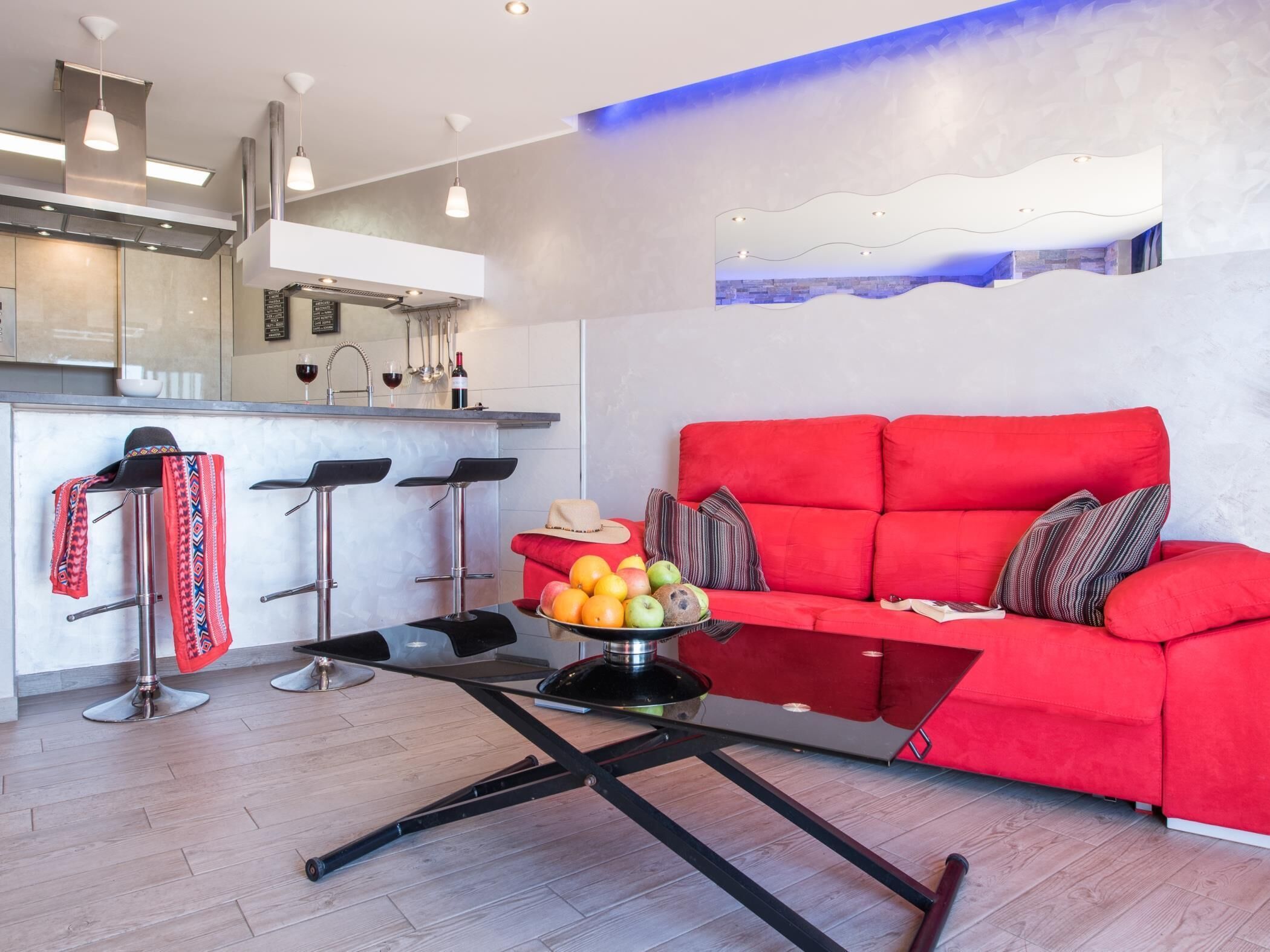 A modern living room with a red sofa, a coffee table with fruits, and a kitchen area with bar stools and a wine bottle.