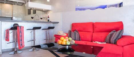 A modern living room with a red sofa, a coffee table with fruits, and a kitchen area with bar stools and a wine bottle.