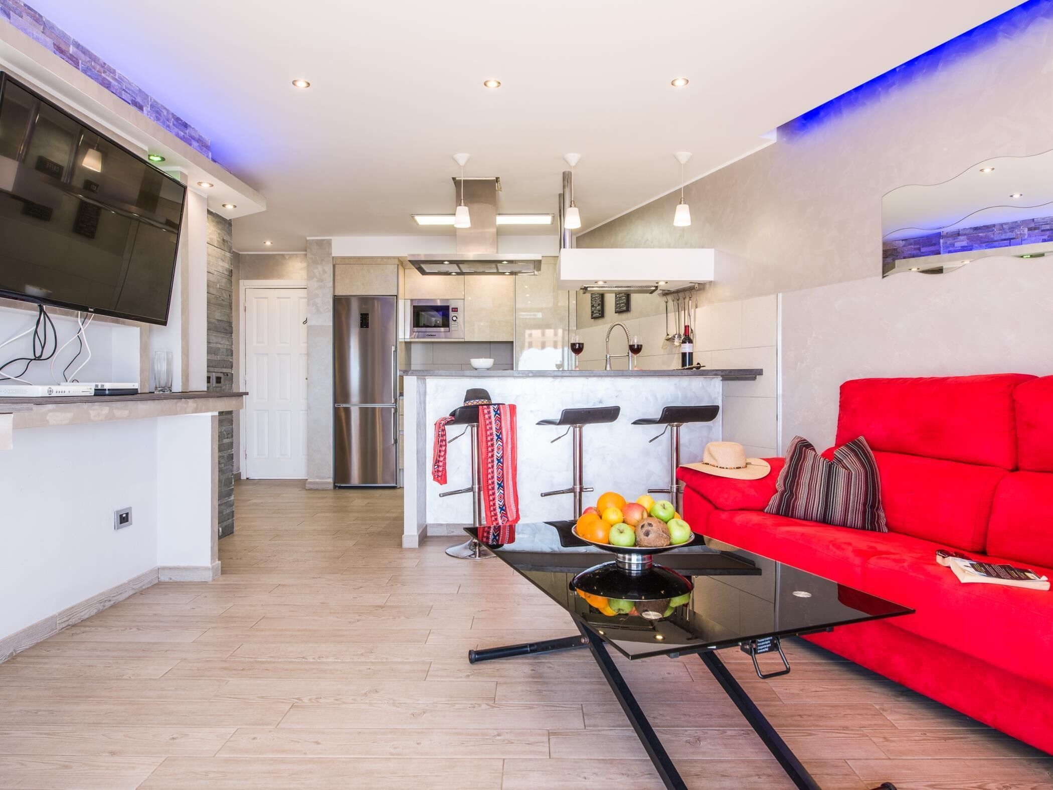 A modern living room with a red sofa, a glass coffee table with fruit, and a kitchen in the background.