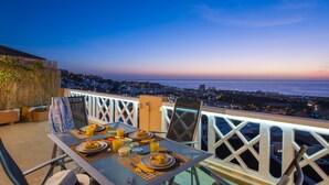 Mesa de jantar posta com itens de café da manhã, com vista para um horizonte costeiro ao entardecer.